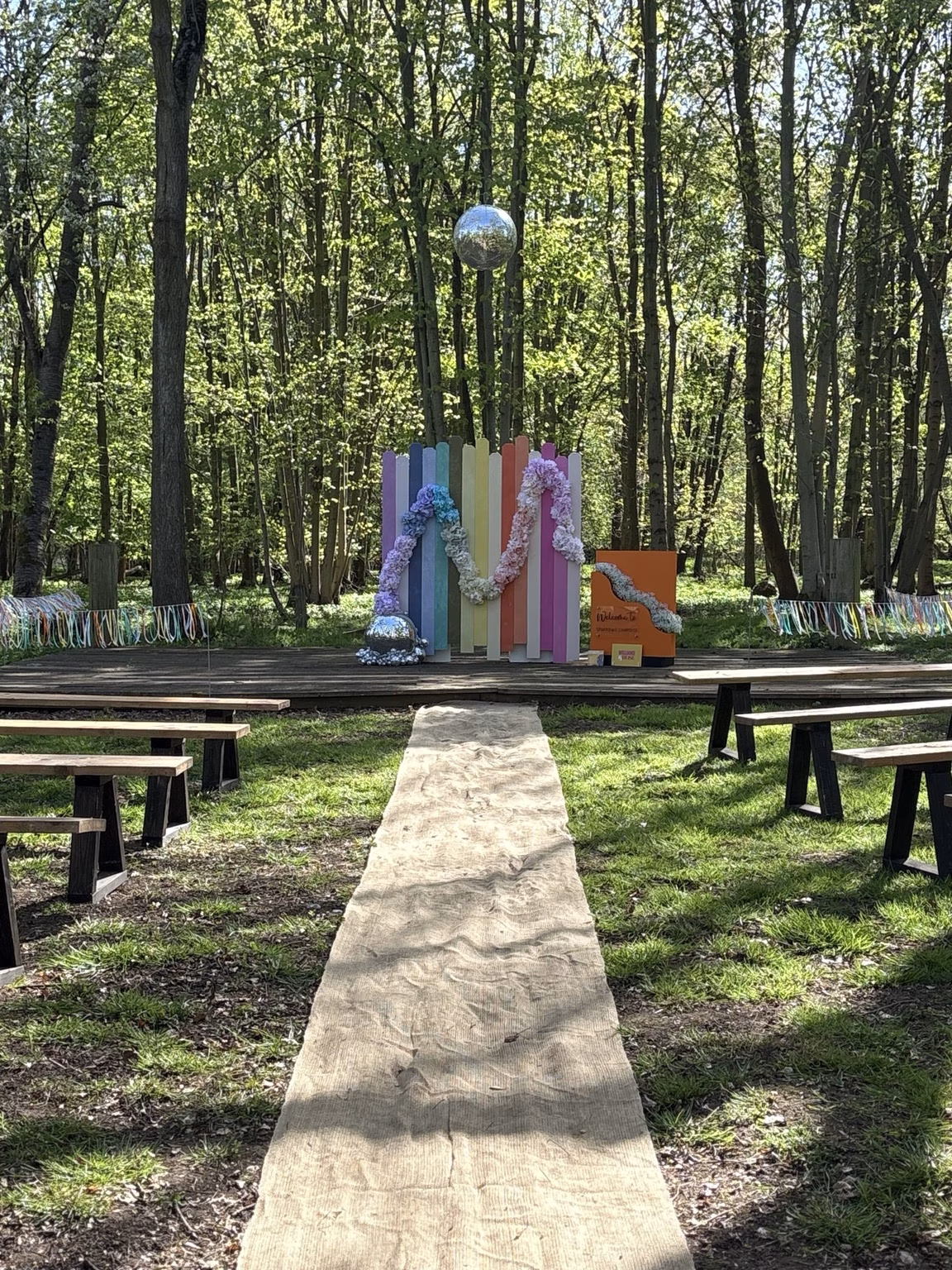 Woodland wedding ceremony area with benches, a pastel plank wall, floral styling and hanging Studio 54 disco balls at Sparrows Campsite.