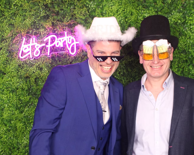 Two wedding guests posing in front of the Fern Wall and Let’s Party neon at Childerley Barn.